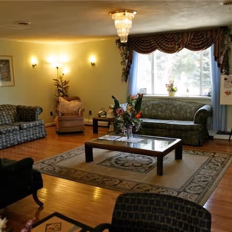 A cozy living room with wooden flooring, featuring a patterned area rug, a glass-top coffee table with a floral arrangement, two sofas, an armchair, a floor lamp, and a large window with decorative curtains letting in natural light.