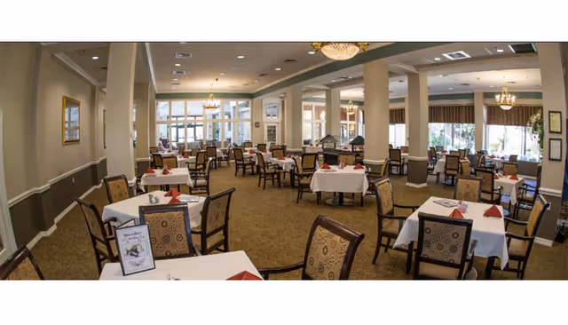 Spacious dining room with multiple tables set with white tablecloths and chairs, chandeliers, and large windows.