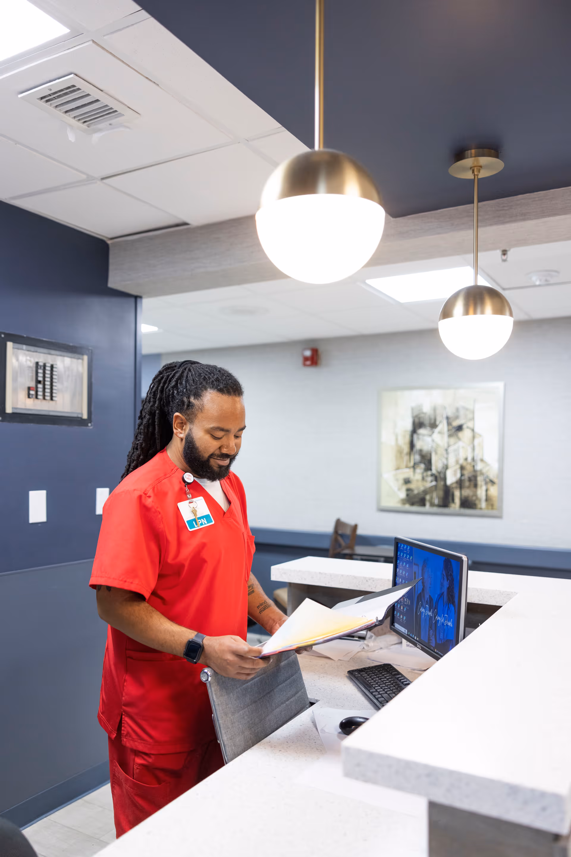 A healthcare worker in red scrubs with an LPN badge is standing at a reception desk, reviewing documents. The desk has a computer monitor, keyboard, and mouse. The background shows a modern interior with hanging round pendant lights and a framed abstract artwork on the wall.