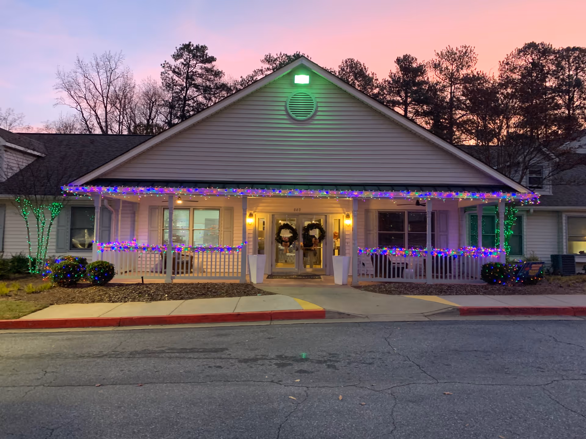 Front exterior view of a single-story building at dusk with holiday lights decorating the porch railing and trees. The building has a peaked roof with a green light at the top and two wreaths on the glass entrance doors. Trees and bushes surround the building, and the sky is a soft pink and purple.