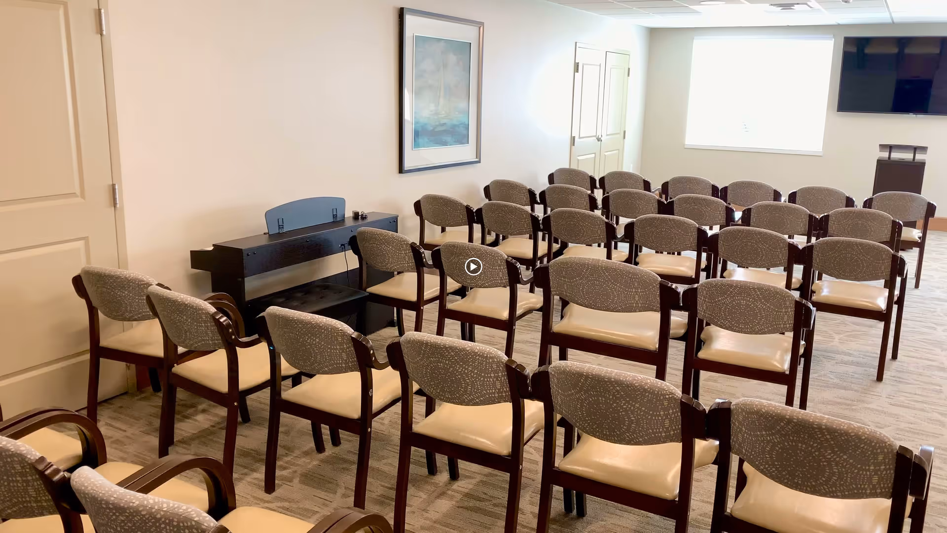 A small conference or meeting room with multiple rows of wooden chairs with cushioned seats and backs arranged facing a podium and a wall-mounted TV. There is a digital piano against the wall with a framed picture above it, and a window letting in natural light.