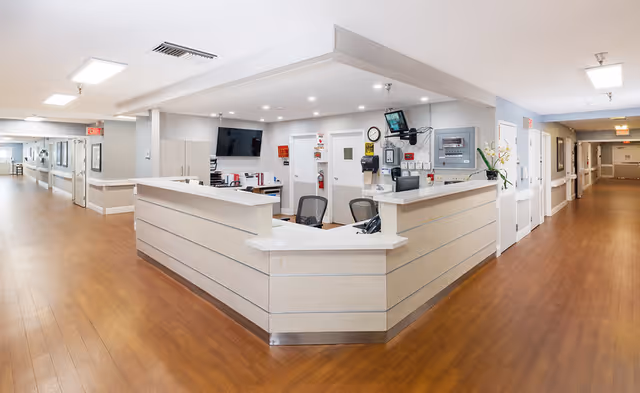 A clean and well-lit nursing station in a senior living facility with a light wood reception desk, two office chairs, a wall-mounted TV, and various medical and office equipment. The station is situated in a wide hallway with wood flooring and white walls, leading to multiple rooms and exits.