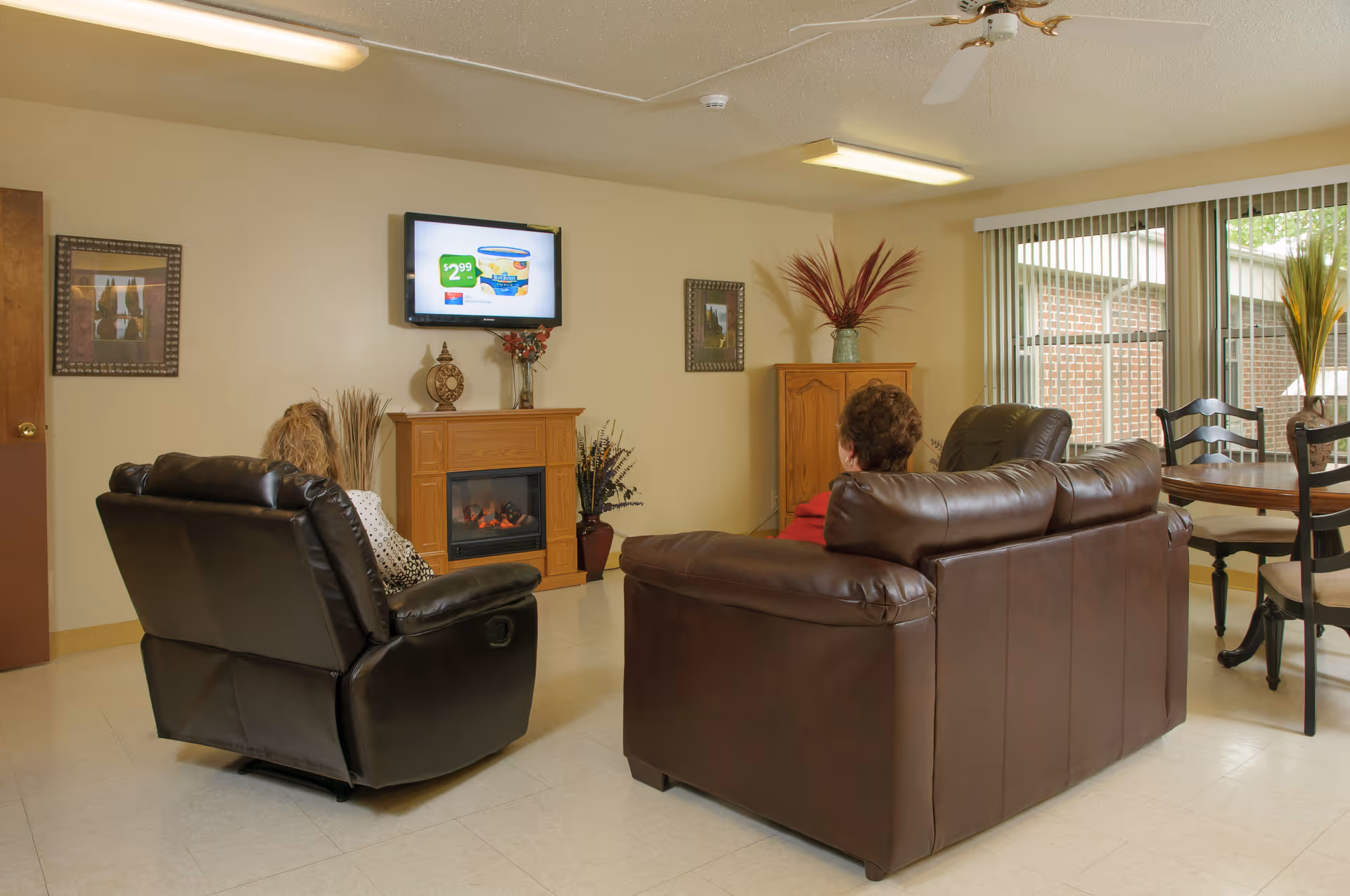 A cozy living room area with two people seated on brown leather furniture facing a wall-mounted TV above a wooden electric fireplace. The room has beige walls, framed pictures, decorative plants, a ceiling fan, and a round wooden dining table with chairs near a large window with vertical blinds.