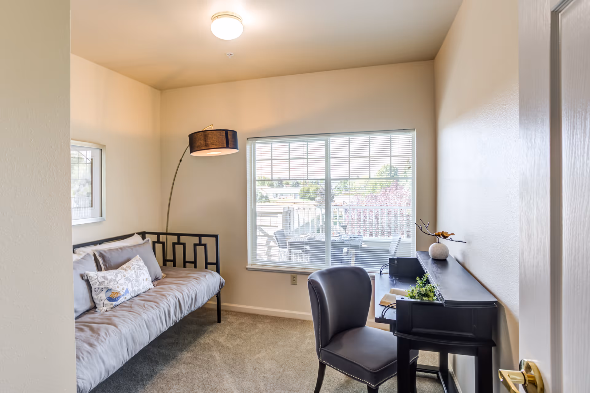 A small, bright room with beige walls and carpeted floor featuring a black metal-framed daybed with gray cushions and decorative pillows on the left side. A tall floor lamp with a black shade stands behind the daybed. On the right side, there is a black desk with a matching chair, a small plant, a decorative vase, and some books. A large window with blinds lets in natural light and shows an outdoor patio area with chairs and a table.