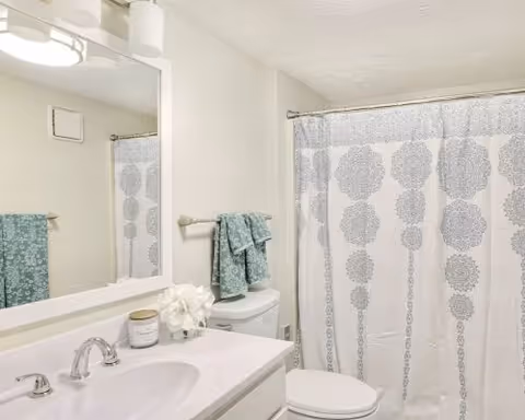 A clean bathroom with a white sink and countertop, a silver faucet, a large mirror above the sink, a toilet with folded teal towels on a towel rack above it, and a shower with a white curtain featuring a gray decorative pattern.