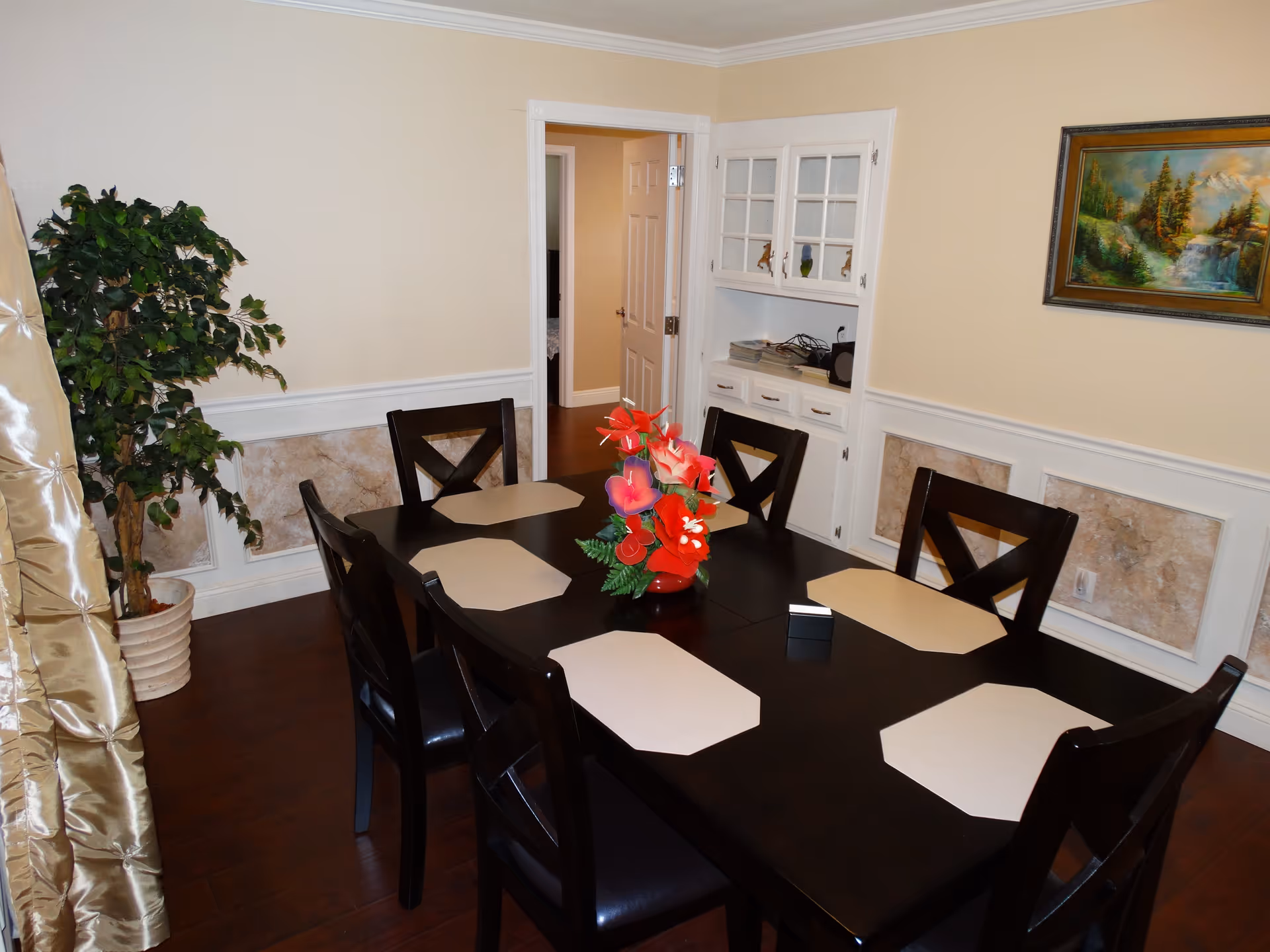 A dining room with a dark wooden table set with six chairs. The table has beige placemats and a red vase with colorful artificial flowers in the center. There is a potted plant in the corner, a built-in white cabinet with glass doors, and a landscape painting on the wall. The floor is wooden, and there is a doorway leading to another room.
