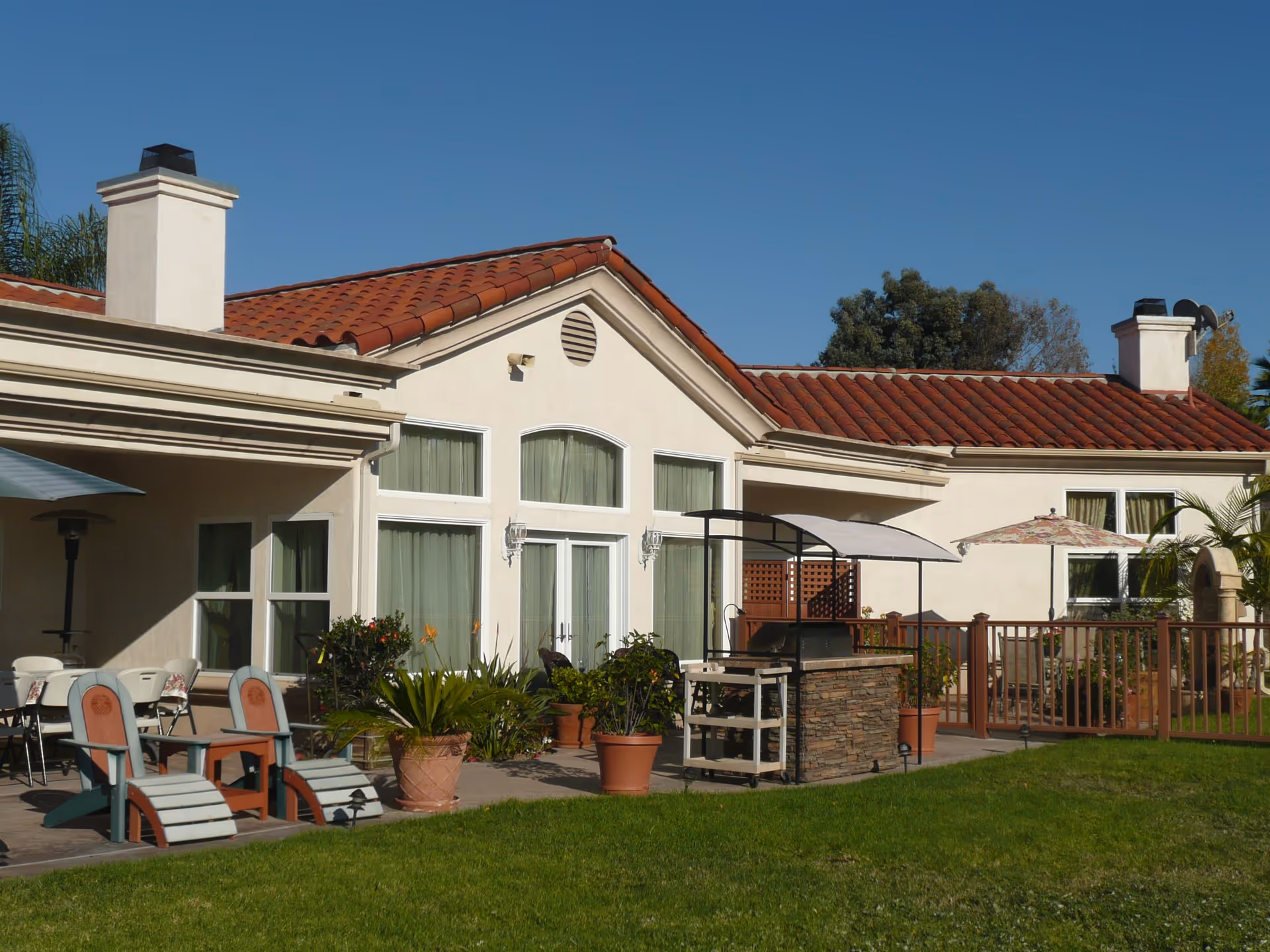 Outdoor patio area of a senior living facility with beige walls and red tile roof. The patio features several chairs, tables with umbrellas, potted plants, and a stone barbecue grill. The sky is clear and blue, and there is a grassy lawn in the foreground.