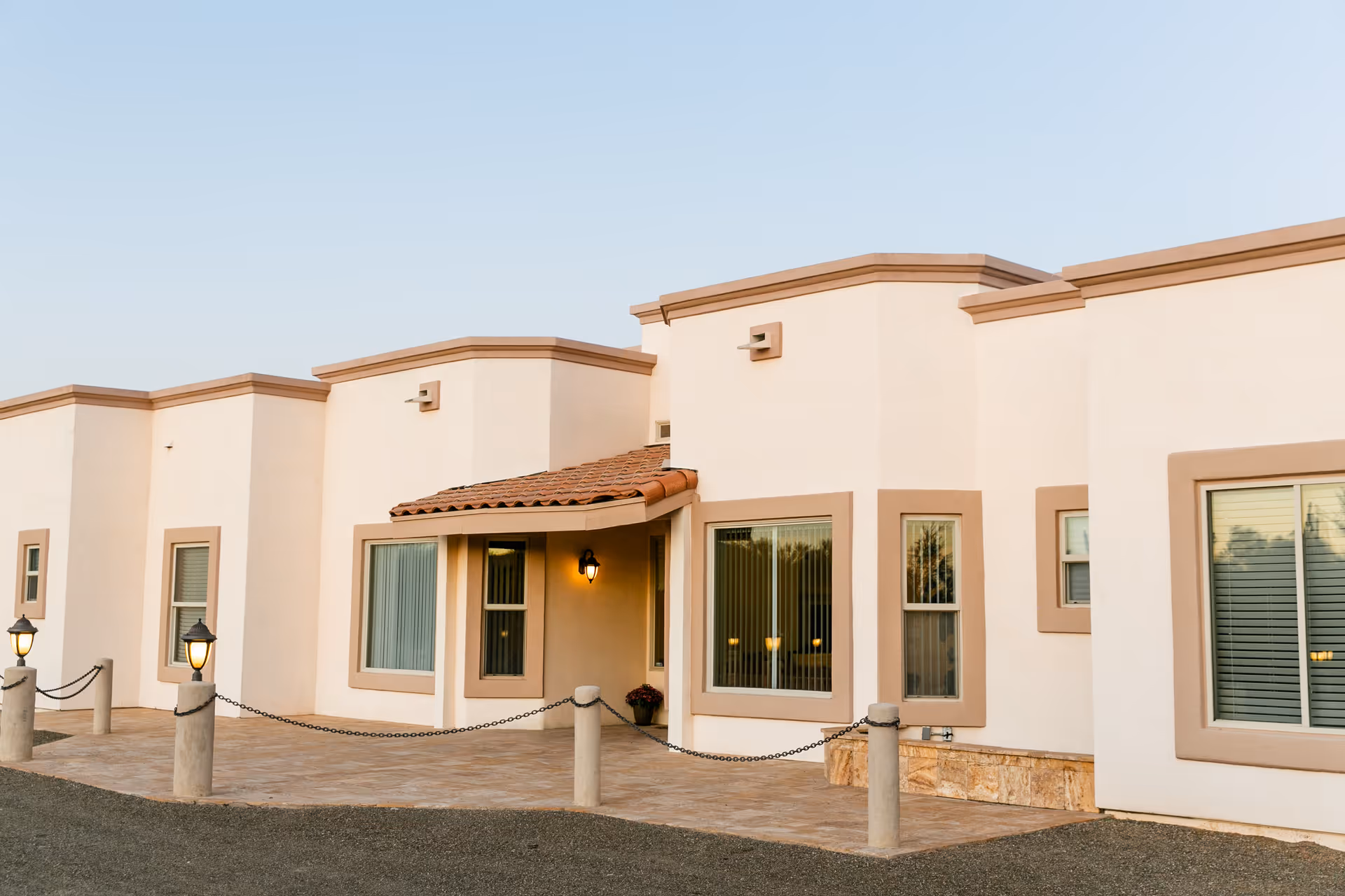 Exterior view of a single-story building with beige walls and brown trim, featuring several windows with blinds and a small covered entrance with a tiled roof and a lit wall-mounted lantern. The area in front has a paved walkway bordered by short concrete posts connected with chains, and two lamp posts with lights on either side.