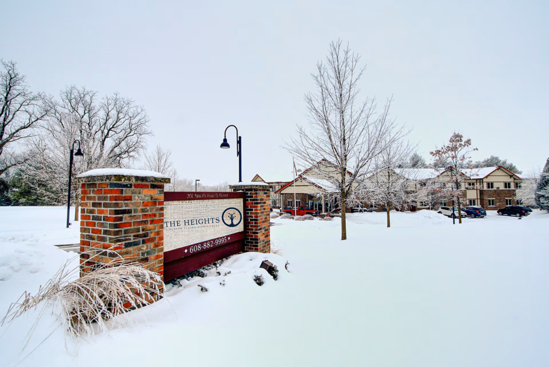 Snow-covered entrance sign for The Heights at Evansville Manor with a brick base and lamp posts, surrounded by snow-covered trees and a building in the background.