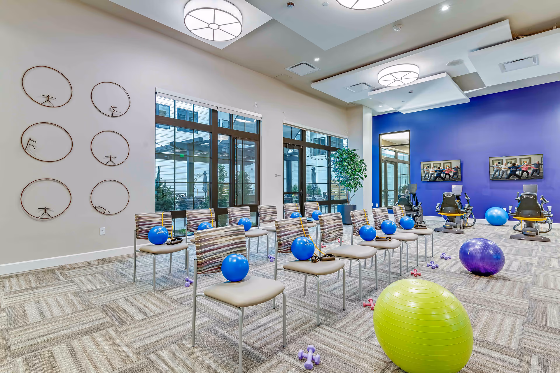 A bright exercise room with rows of chairs, each holding a blue exercise ball and resistance bands. The room has large windows letting in natural light, a blue accent wall with two televisions showing a group exercise class, and various exercise equipment including stationary bikes, dumbbells, and large exercise balls on the carpeted floor.