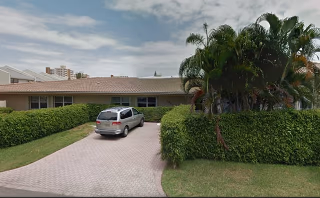 Single-story building with a tiled roof partially obscured by green hedges and palm trees, a silver minivan parked on a brick driveway under a partly cloudy sky.