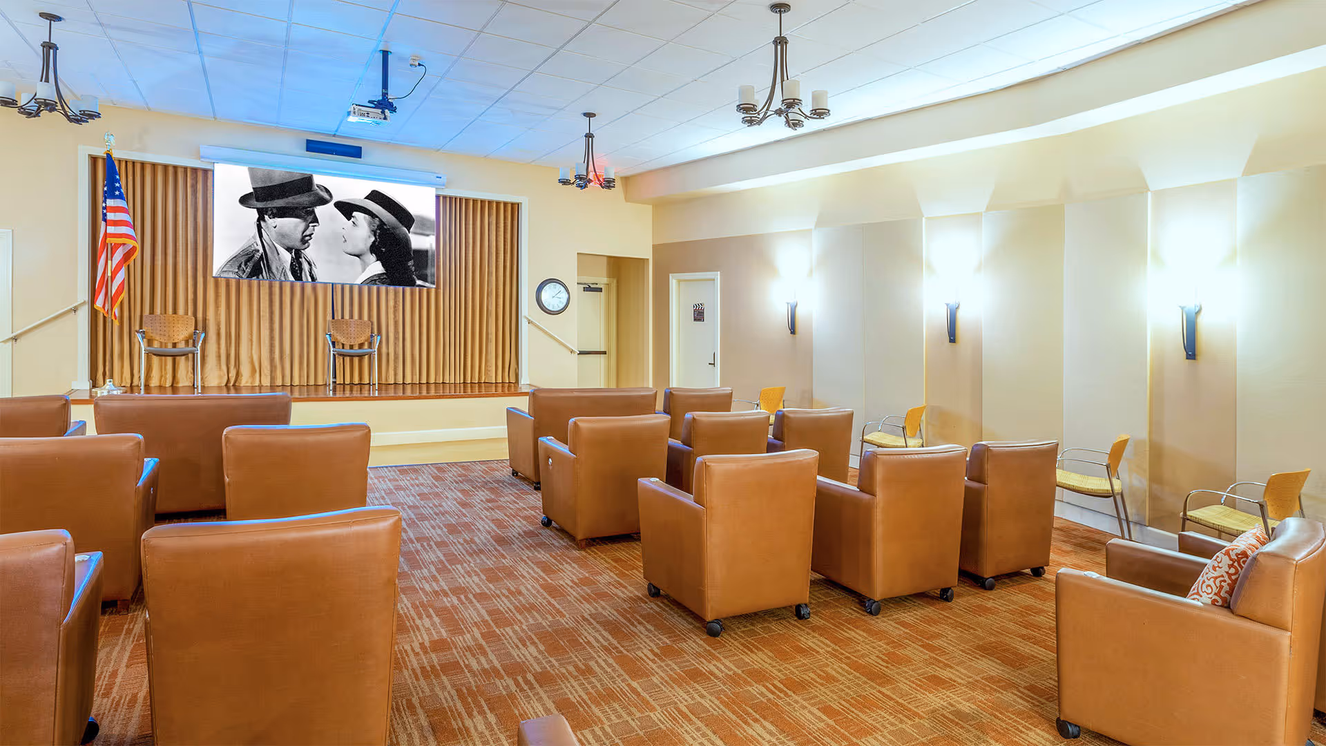 A small theater room with brown leather armchairs arranged in rows facing a stage with a projector screen showing a black and white movie scene. The room has beige walls, wall sconces for lighting, an American flag on the left side of the stage, and a clock on the wall near a door.