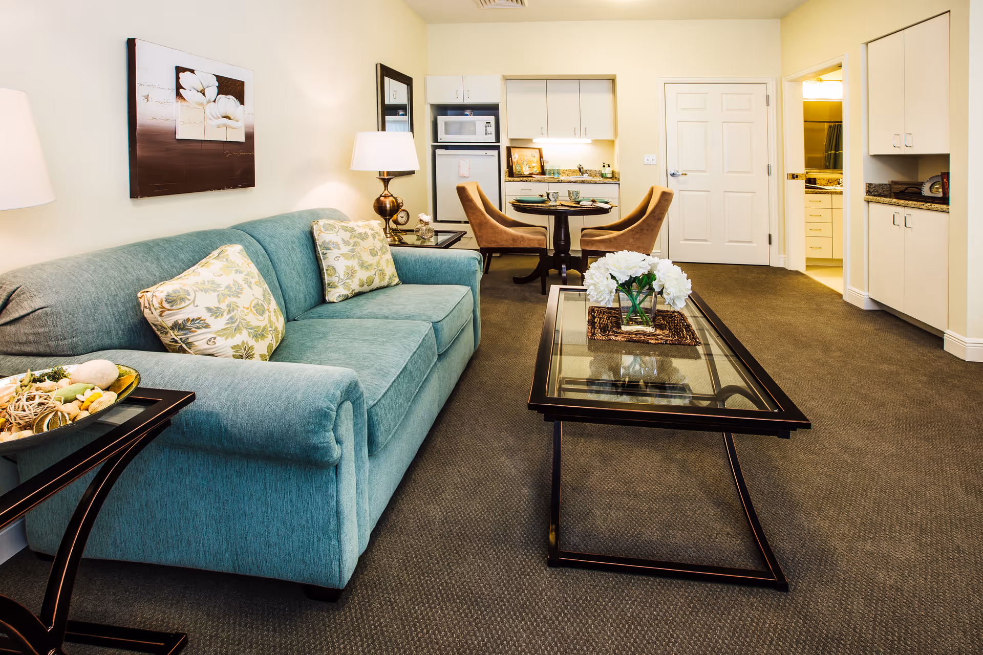 A cozy senior living room featuring a teal sofa with floral cushions, a glass coffee table with a vase of white flowers, a side table with decorative items, and a small dining area with two chairs and a round table. The background shows a kitchenette with white cabinets and a microwave, and a doorway leading to a bathroom.