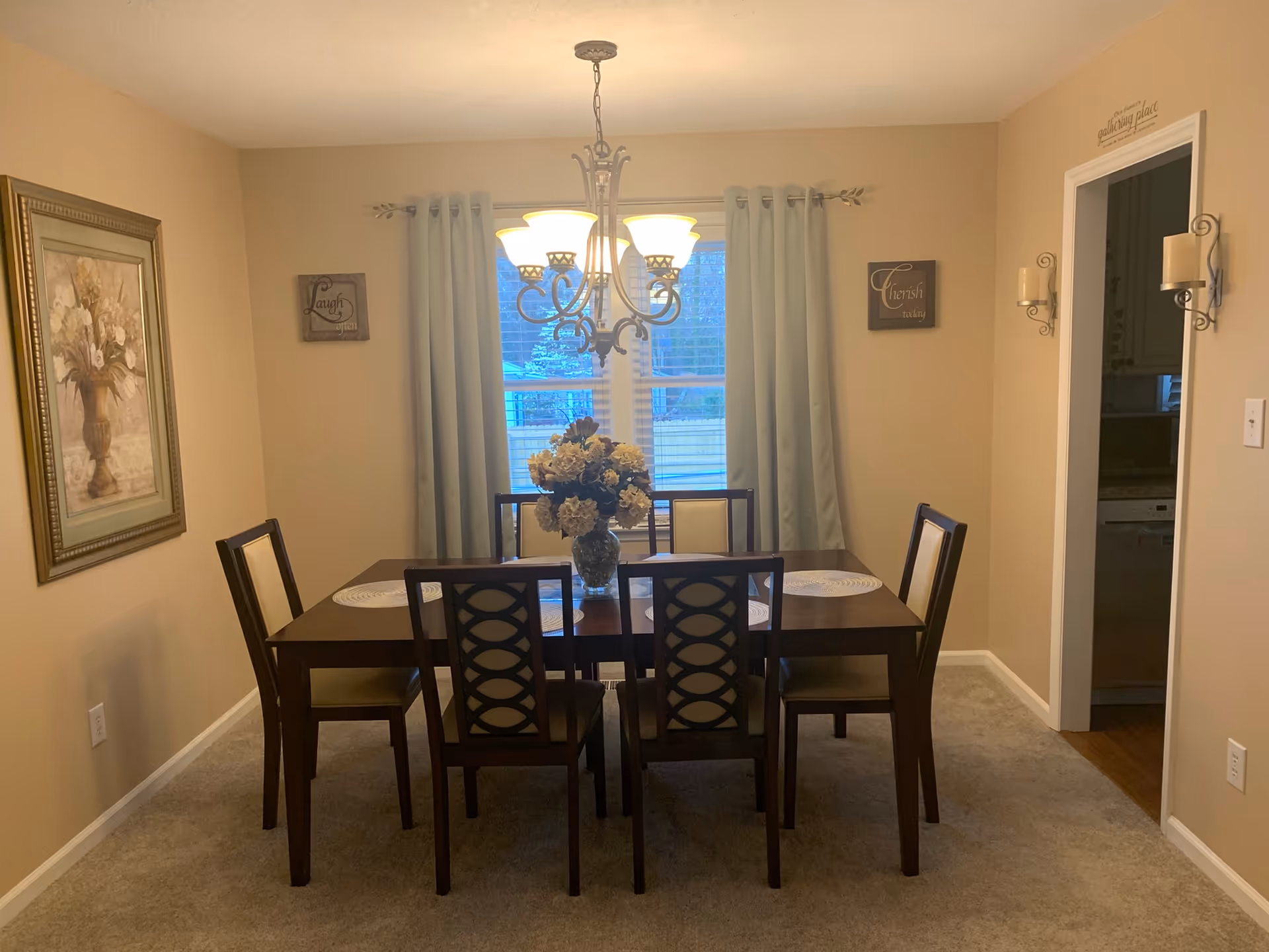 A dining room with a rectangular wooden table and six chairs. A vase with flowers is placed in the center of the table. The room has beige walls, a window with light blue curtains, and a chandelier hanging above the table. There are framed artworks and decorative wall plaques on the walls. A doorway leads to a kitchen area.