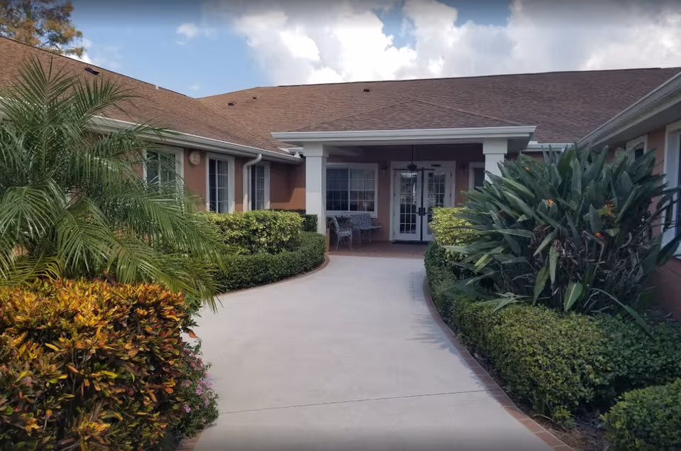 Entrance walkway to a single-story building with a covered porch area, surrounded by well-maintained bushes and tropical plants under a partly cloudy sky.
