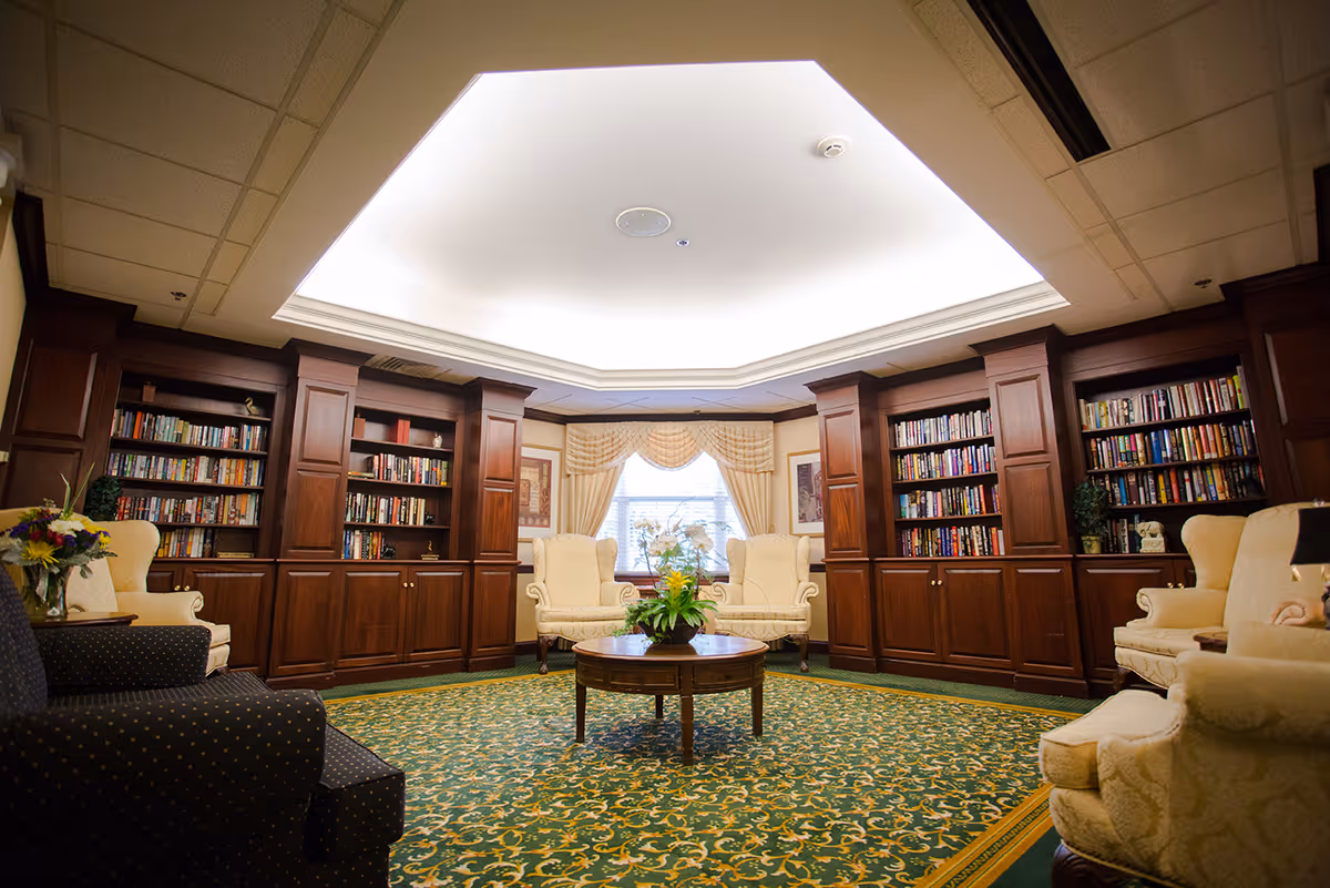 A cozy library or sitting room with built-in wooden bookshelves filled with books, several upholstered armchairs arranged around a round wooden coffee table with a flower arrangement, and a large window with draped curtains letting in natural light.