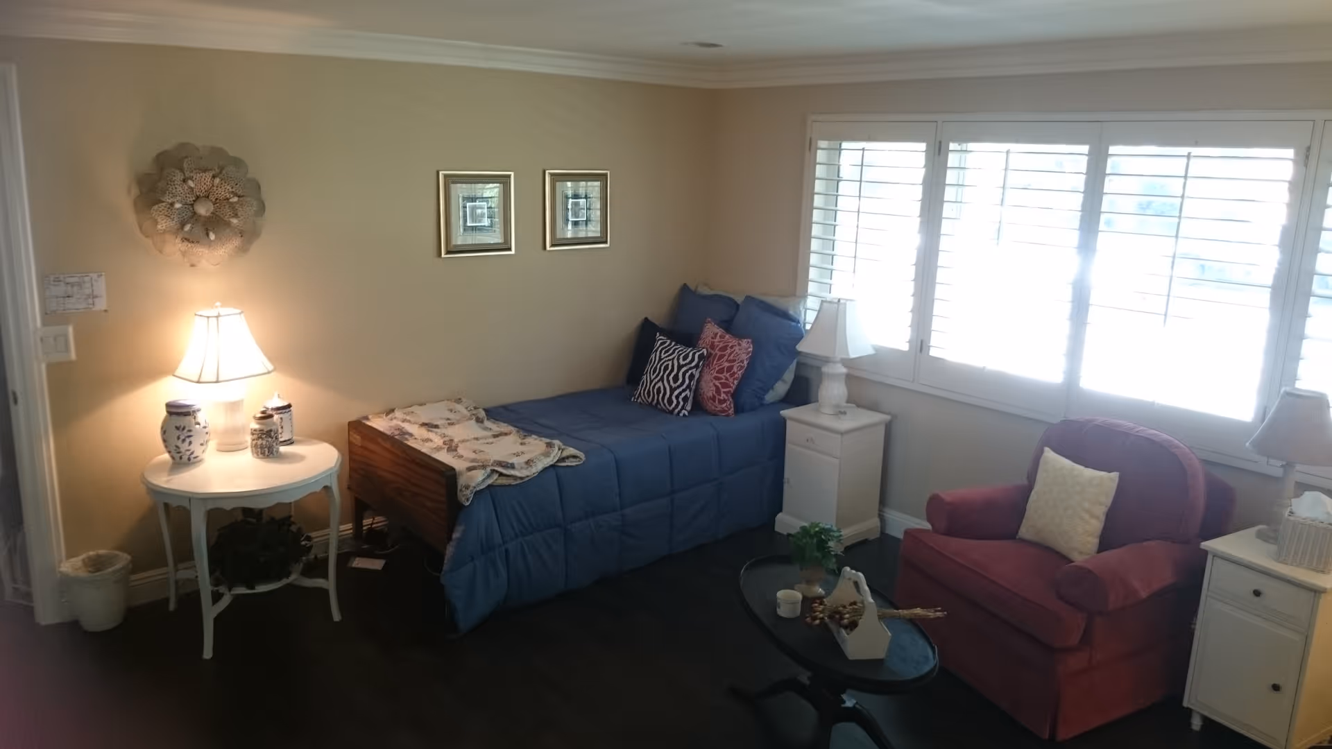 A cozy bedroom with a single bed covered in a blue comforter and several decorative pillows. Next to the bed is a white nightstand with a lamp, and a small round table with another lamp and decorative items. A red armchair with a light-colored pillow sits near a window with white shutters. The room has beige walls and dark wood flooring.