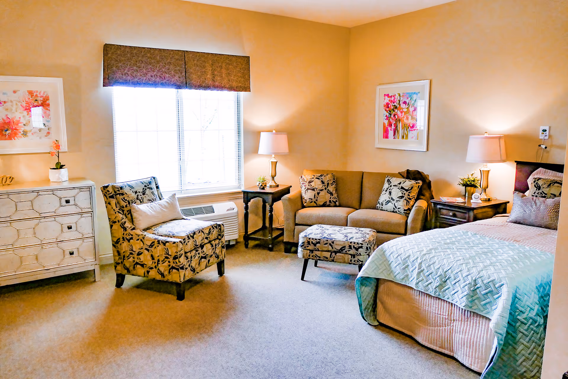 A cozy bedroom in the Inn at Bear Trail featuring a bed with a light blue quilt, a beige sofa with patterned cushions, a matching patterned armchair and ottoman, two side tables with lamps, a window with a valance, and floral artwork on the walls.