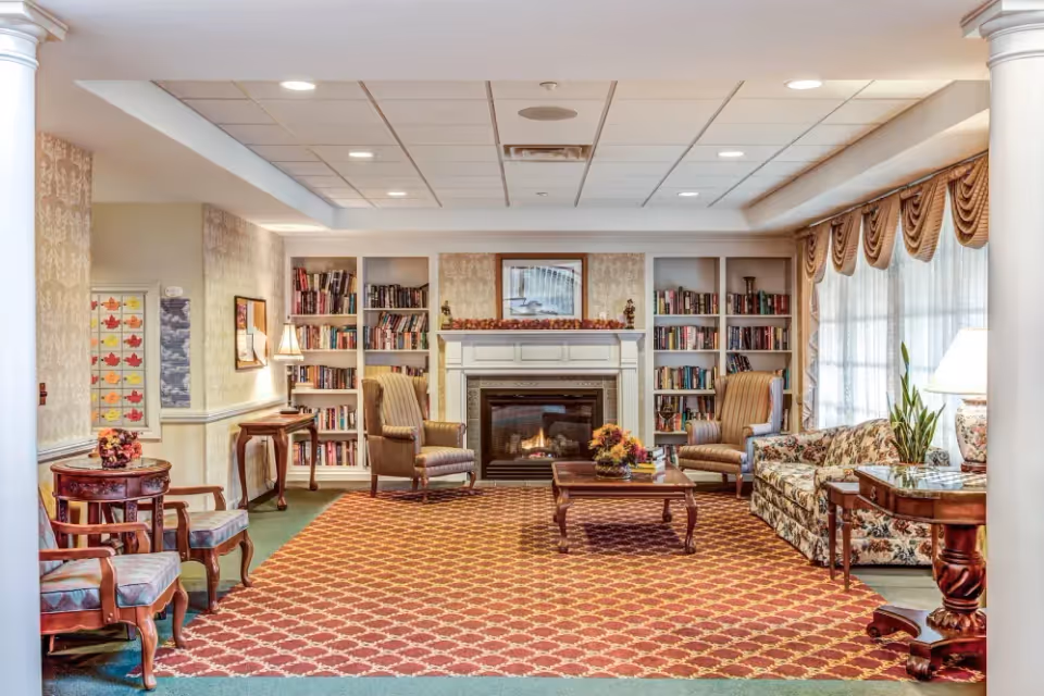 A cozy living room area with a patterned carpet, two striped armchairs, a floral sofa, and a wooden coffee table with a flower arrangement. The room features a fireplace with a mantel decorated with autumn leaves and a framed picture above it. Built-in bookshelves filled with books flank the fireplace. Large windows with sheer curtains and valances allow natural light to enter. There are also side tables with lamps and plants, and decorative columns framing the space.