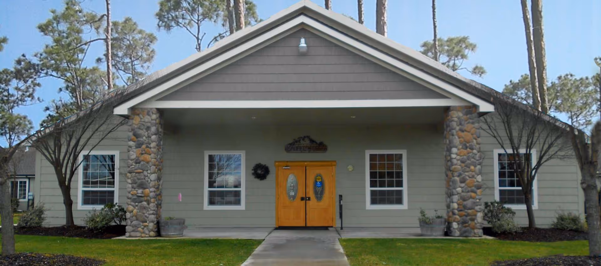 Front exterior view of a single-story building with a peaked roof, two stone pillars flanking a double wooden door with oval glass inserts, two windows on either side, small trees and shrubs in front, and a concrete walkway leading to the entrance under a covered porch.