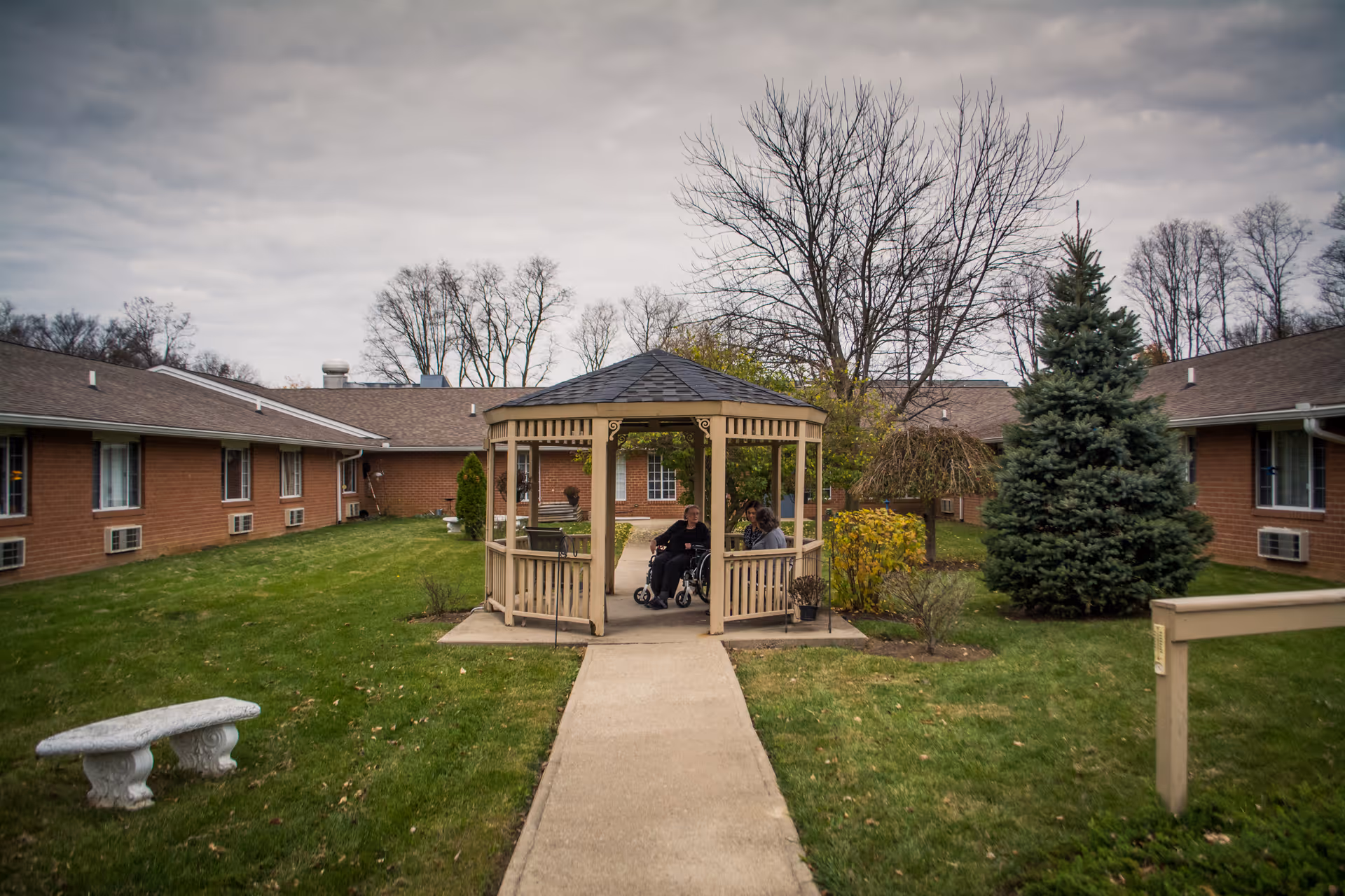 Outdoor courtyard area of a senior living facility with a gazebo in the center. Two people are sitting inside the gazebo, one in a wheelchair. The courtyard is surrounded by single-story brick buildings with windows and air conditioning units. There is a concrete pathway leading to the gazebo, green grass, trees without leaves, and a cloudy sky overhead.