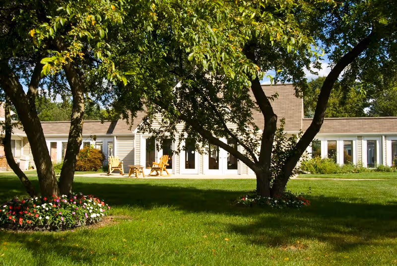 View of a single-story building with large windows and a patio area with wooden chairs, surrounded by green grass, trees, and flower beds under a clear blue sky.