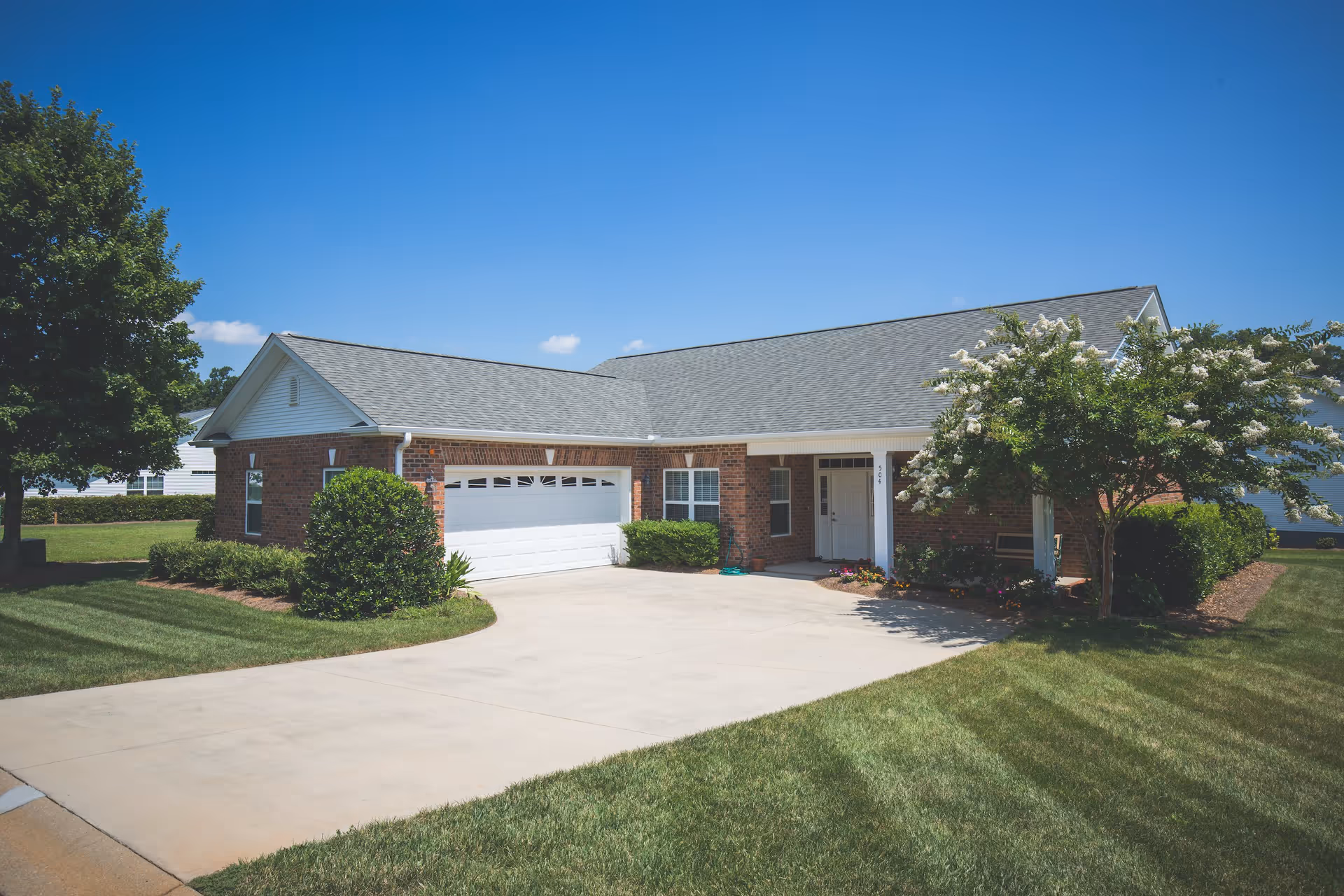 Exterior view of a single-story brick house with a two-car garage, a concrete driveway, and well-maintained green lawn and shrubs under a clear blue sky.