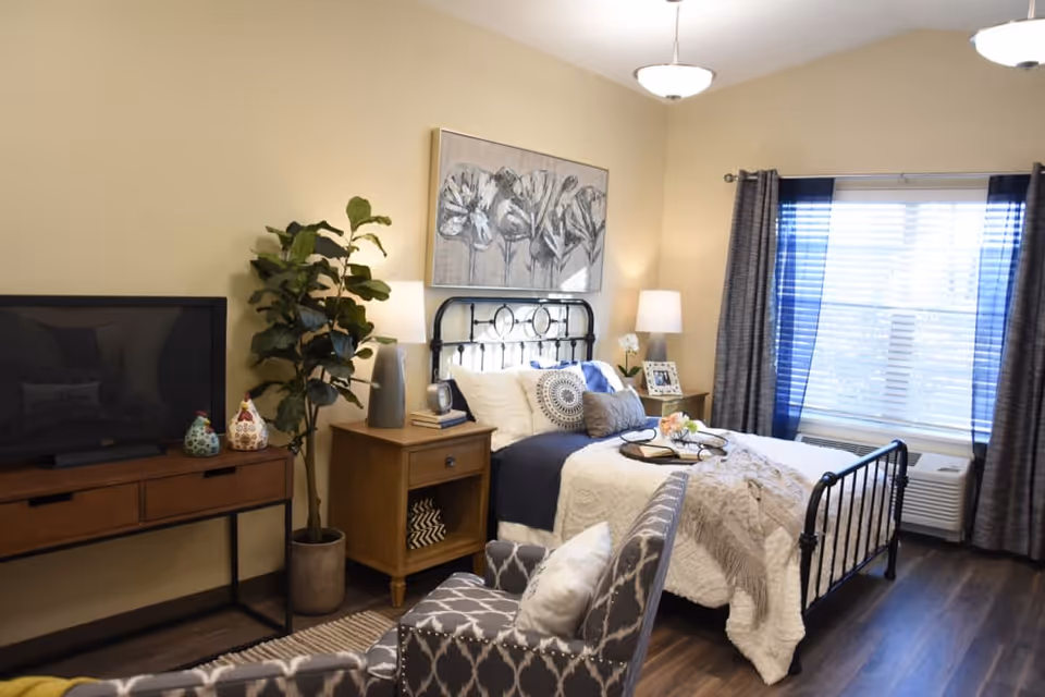 A cozy furnished bedroom with a black metal bed, bedside tables and lamps, a TV on a console, an armchair, and a window with curtains.