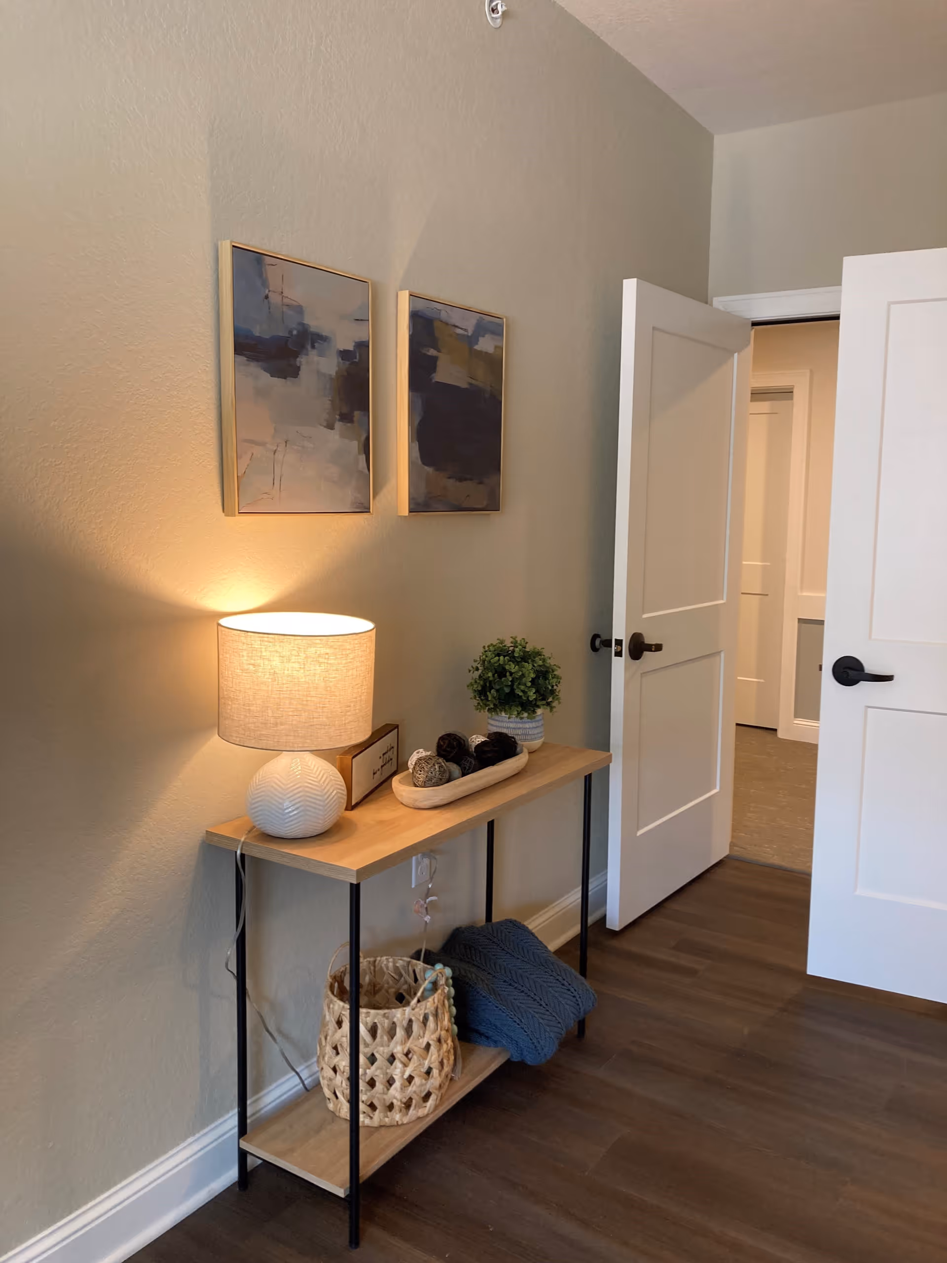 A small wooden console table with black metal legs against a beige wall. On the table is a white textured lamp with a beige lampshade, a small potted plant, a decorative wooden bowl with yarn balls, and a small framed sign. Below the table is a woven basket and a folded dark blue knitted blanket. Two abstract paintings in wooden frames hang on the wall above the table. A white door is partially open, showing a hallway with similar flooring.
