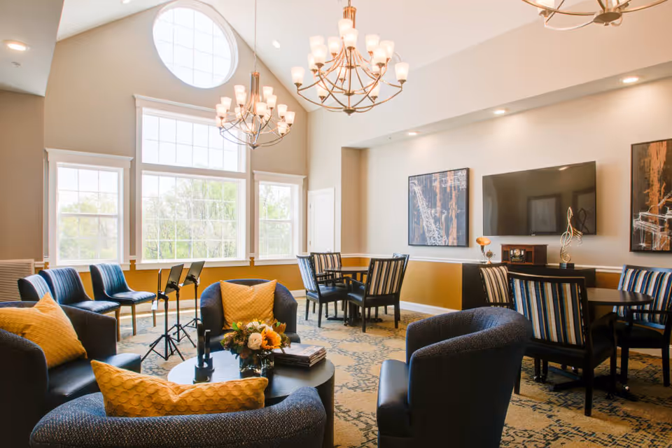 A bright and spacious common area with large windows letting in natural light. The room features comfortable blue armchairs with yellow pillows arranged around a round coffee table with a flower arrangement and books. There are multiple tables with striped chairs, a wall-mounted flat screen TV, and modern chandeliers hanging from a high ceiling with a large circular window above.