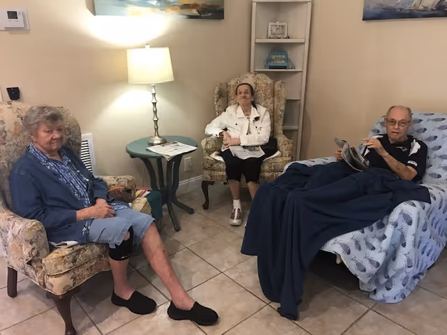 Three elderly individuals sitting in a cozy living room area. Two women are seated on floral armchairs, and a man is reclining on a cushioned chair covered with a blanket. A small round table with a lamp and some papers is between the women. The room has tiled flooring and light-colored walls with framed artwork.