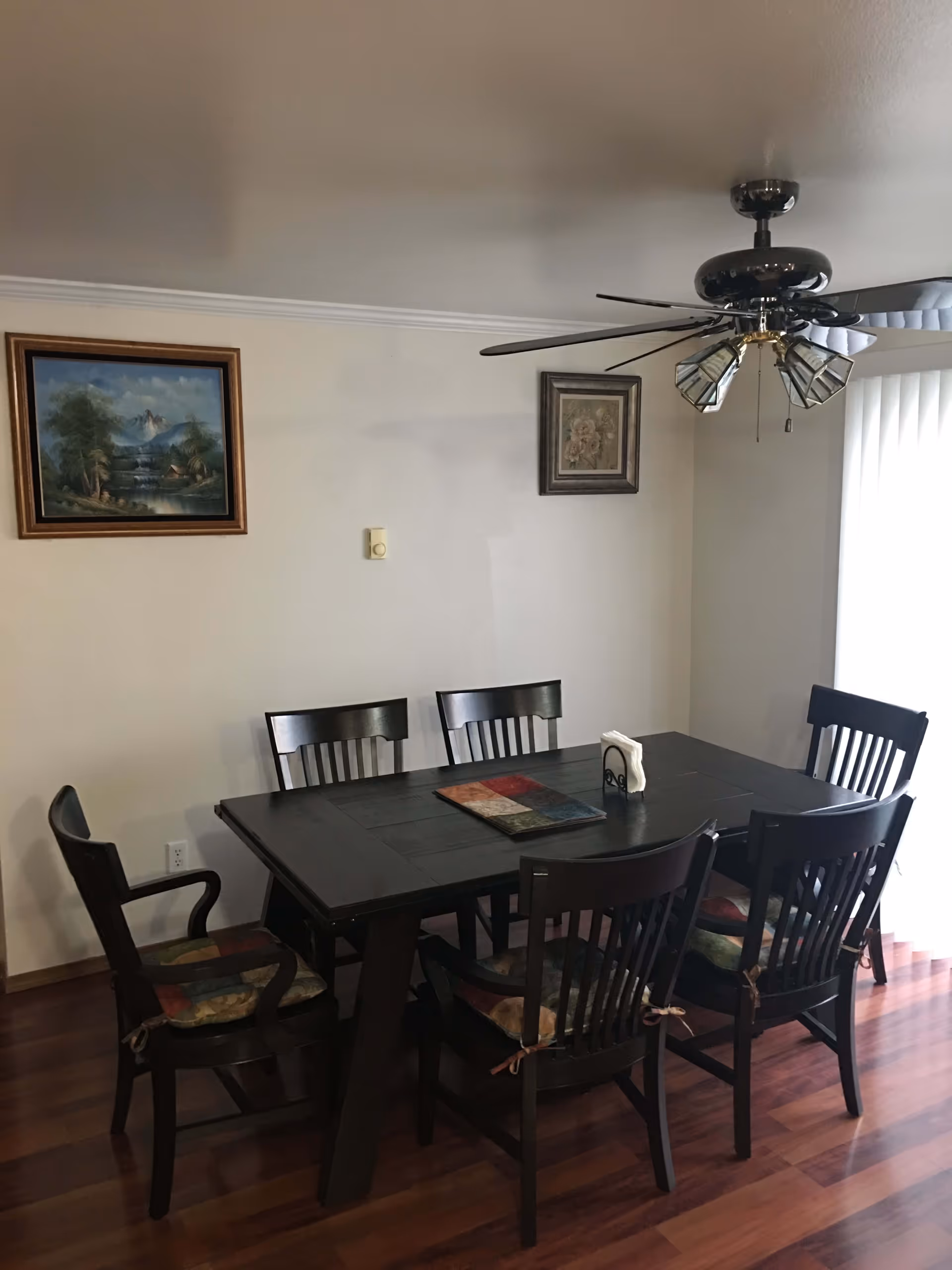 A dining room with a dark wooden rectangular table surrounded by six matching chairs with patterned cushions. The room has wooden flooring, two framed paintings on the wall, a ceiling fan with lights, and vertical blinds covering a window or sliding door.