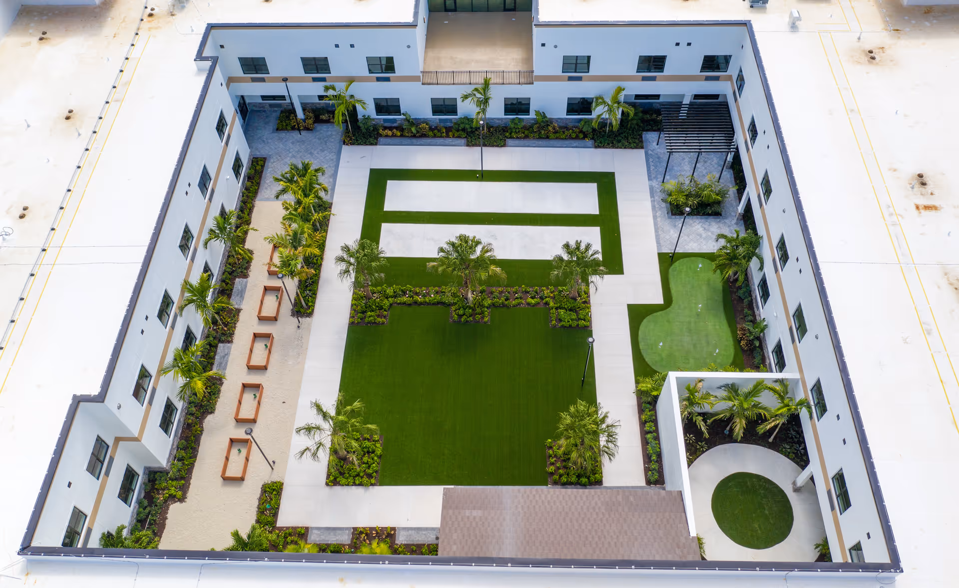 Aerial view of a rectangular courtyard surrounded by a white building with multiple windows. The courtyard features neatly manicured green lawns, palm trees, paved walkways, benches, and landscaped garden areas. There is a small putting green area on the right side and a covered circular seating area in the bottom right corner.