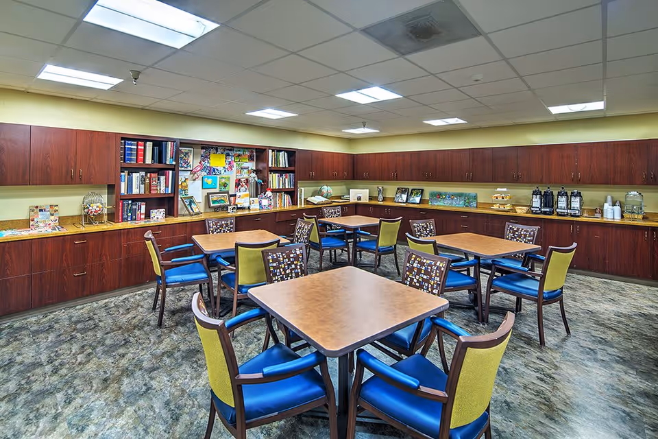 A well-lit room with several square tables surrounded by chairs with blue seats and patterned backs. The room has wooden cabinets lining the walls, with shelves holding books, framed pictures, and decorative items. On the right side, there is a counter with coffee dispensers, cups, and a tiered tray with snacks. The floor has a marbled pattern, and the ceiling has recessed lighting panels.