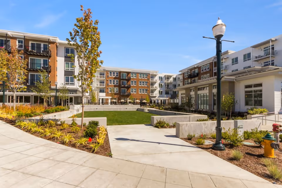Landscaped courtyard and lawn surrounded by multi-story apartment buildings and walkways under a blue sky.