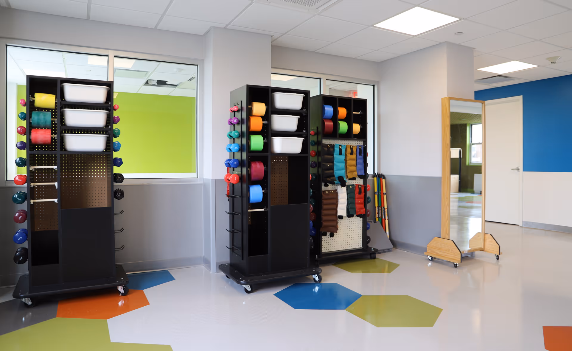 Bright therapy/activity room with storage carts holding colorful exercise equipment and a full-length mirror against a wall.