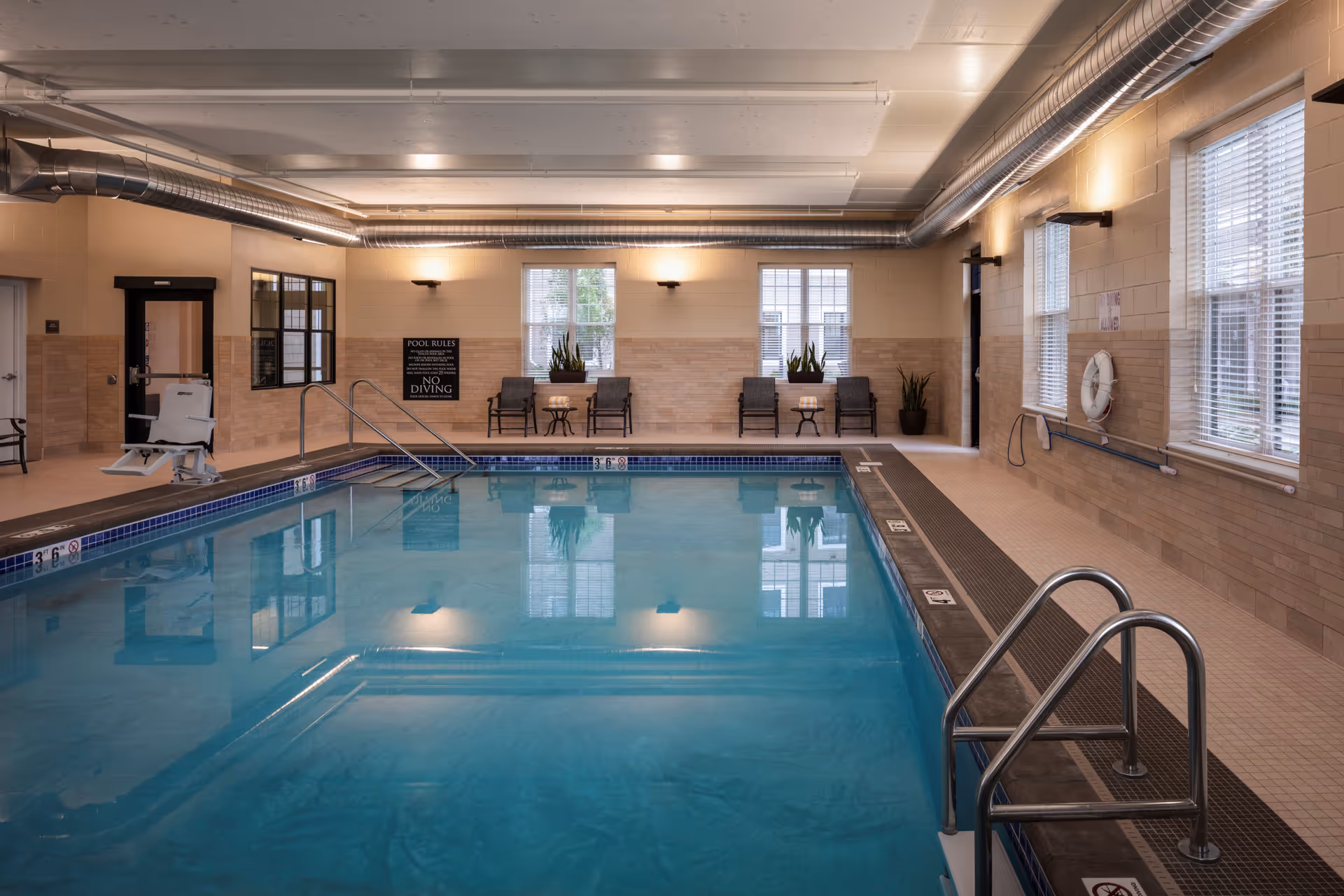 Indoor swimming pool area with clear blue water, metal handrails, pool lift chair, beige tiled walls, windows with blinds, and seating area with chairs and small tables. The room has exposed ductwork on the ceiling and pool safety signs on the walls.