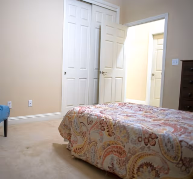 A furnished bedroom with a patterned bedspread, beige walls, and open doorway to a bright hall.