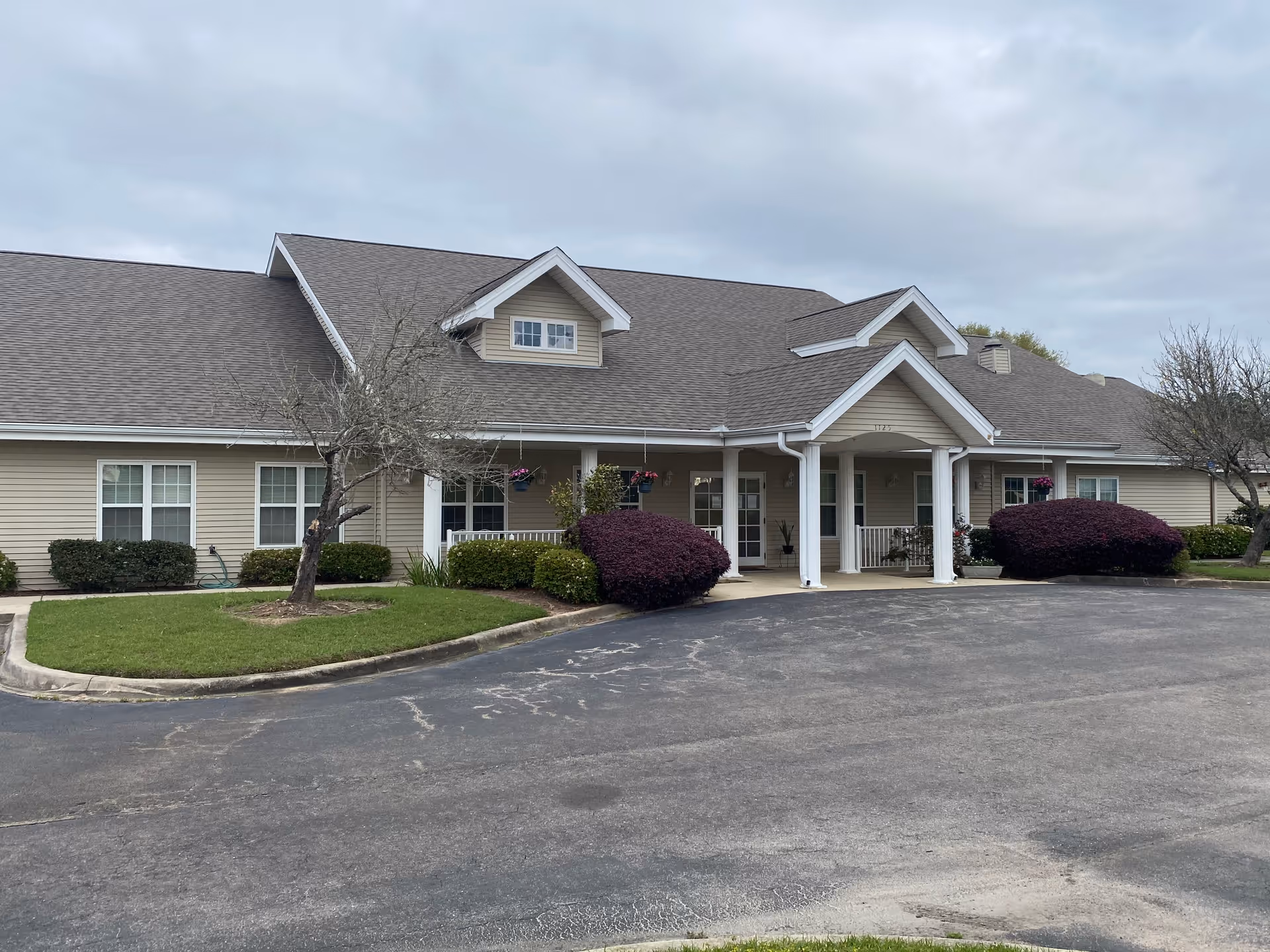 Exterior view of a single-story beige building with a gray shingled roof, white columns at the entrance, and well-maintained landscaping including bushes and small trees under a cloudy sky.