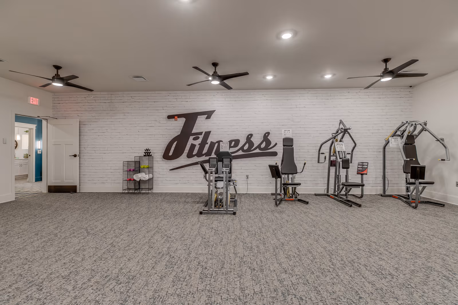 A fitness room with exercise machines lined up against a white brick wall with the word 'Fitness' written in large cursive letters. The room has a carpeted floor, three ceiling fans with lights, and an open door leading to another room.