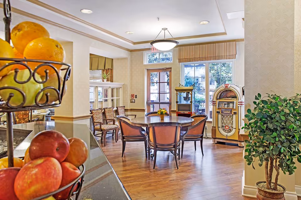 Bright communal dining area with a round table and chairs, a fruit stand in the foreground, and a jukebox and popcorn machine by the windows.