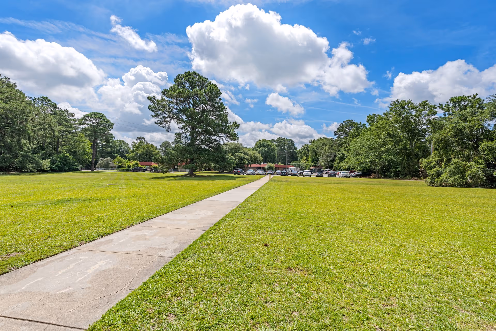 A wide concrete walkway leads through a large, well-maintained grassy area with trees on both sides under a partly cloudy blue sky. In the distance, there are parked cars and a low building with a red roof.