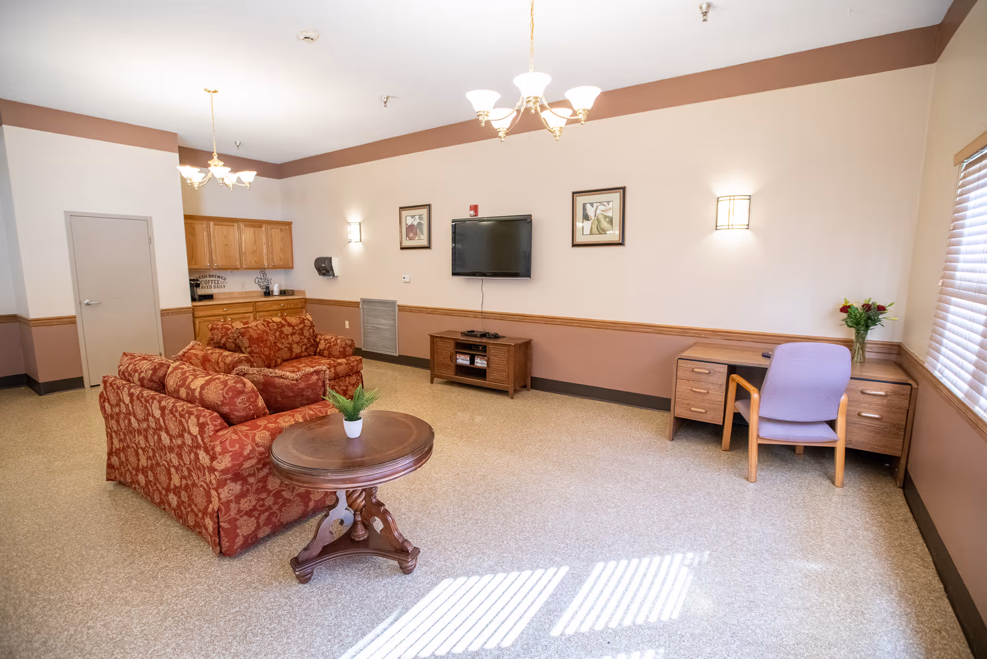 A spacious senior living lounge with red patterned sofas, a round wooden table, wall-mounted TV, desk and cabinets.