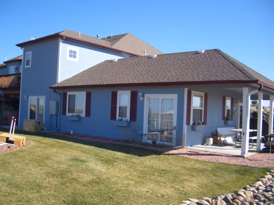 Exterior view of a two-story residential building with light blue siding, brown roof shingles, and maroon window shutters. The building has several windows, a sliding glass door, and a covered patio area with outdoor furniture. The lawn in front is green and well-maintained, with a clear blue sky overhead.