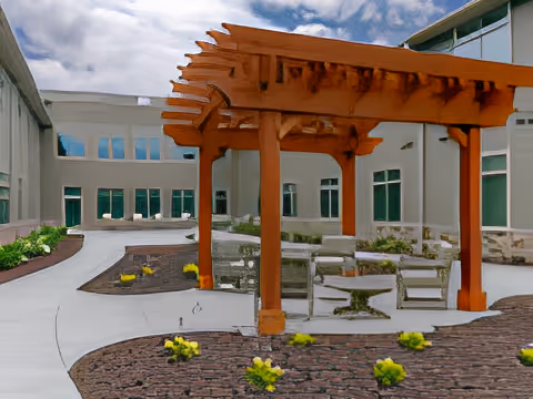 Outdoor courtyard area with a wooden pergola, stone benches, and tables surrounded by landscaped garden beds and a curved concrete walkway, adjacent to a multi-story building with large windows.