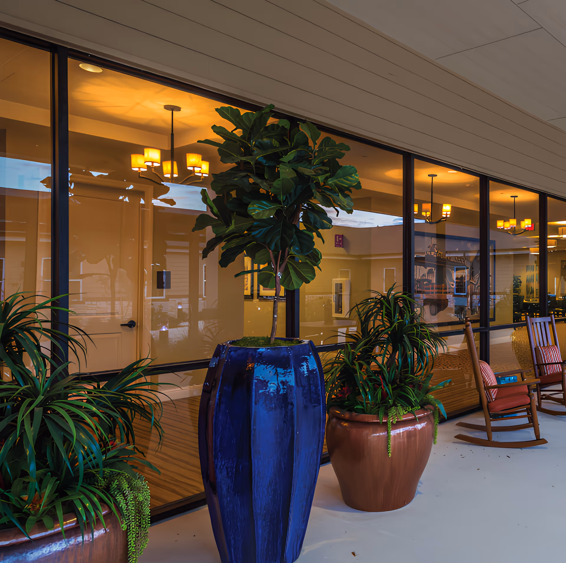 Outdoor covered patio area with large potted plants and wooden rocking chairs with cushions, adjacent to large glass windows showing a warmly lit interior with chandeliers and framed artwork.
