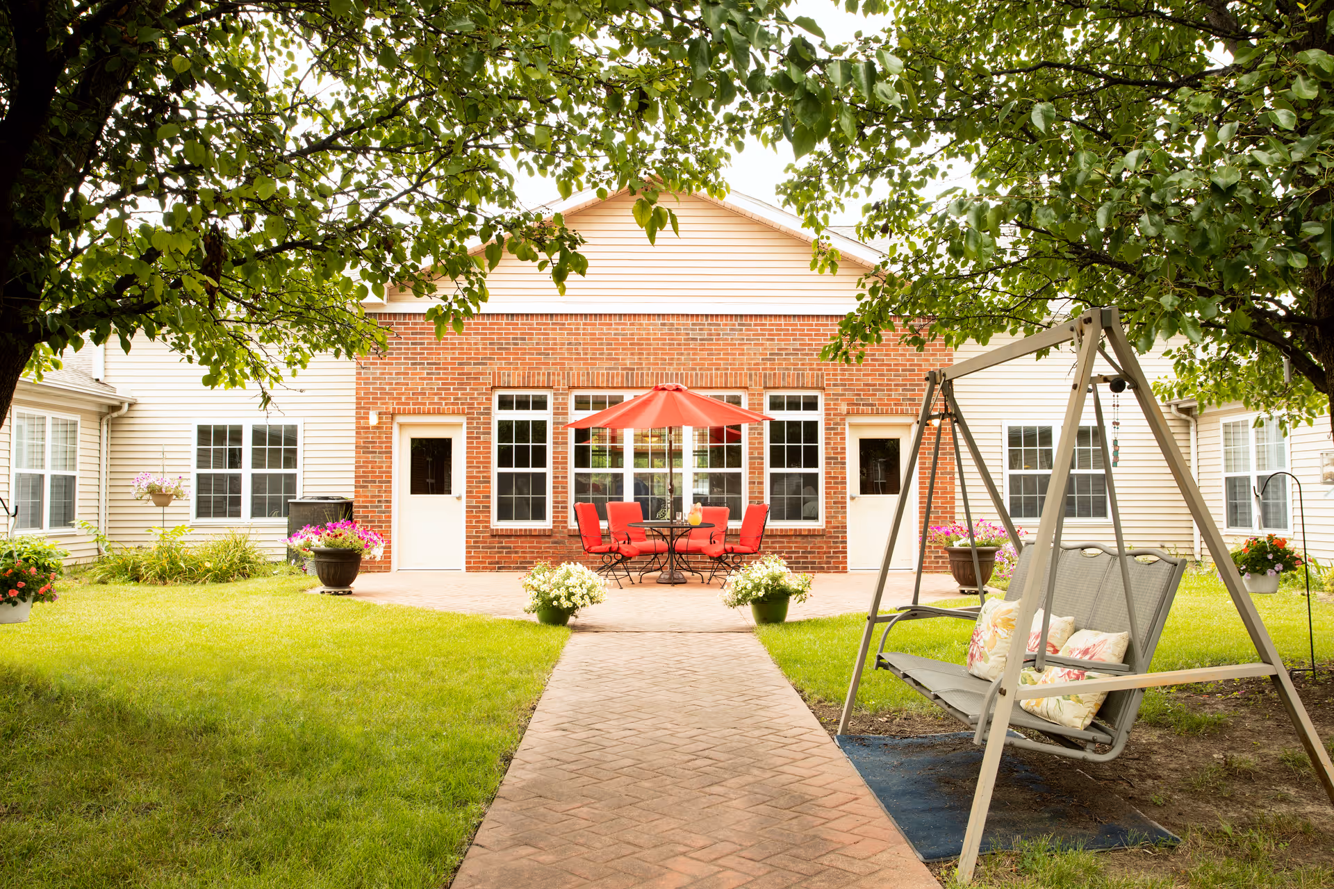 A courtyard with patio furniture under a red umbrella, potted flowers, and a swinging bench in front of a brick-and-siding building.