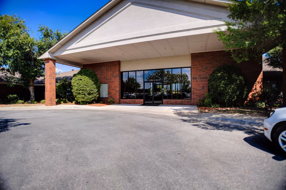 Front exterior view of a building with a covered entrance supported by brick columns, large glass windows and doors, surrounded by trimmed bushes and trees under a clear blue sky.