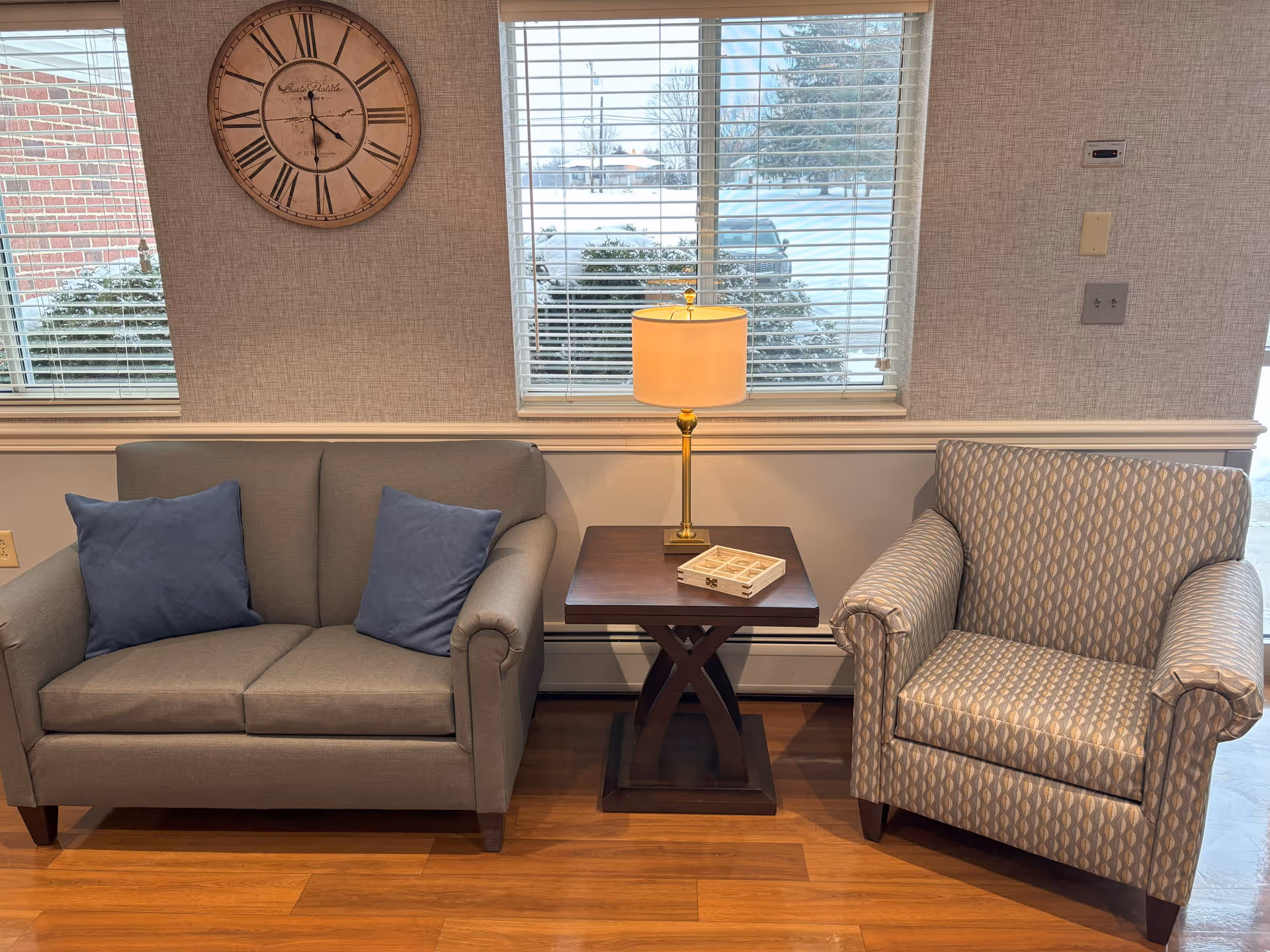A cozy seating area with a gray loveseat featuring two blue pillows on the left, a patterned armchair on the right, and a wooden side table with a lamp and a small decorative box in the center. Behind the seating area are two windows with white blinds, and a large round wall clock is mounted on the wall above the loveseat.