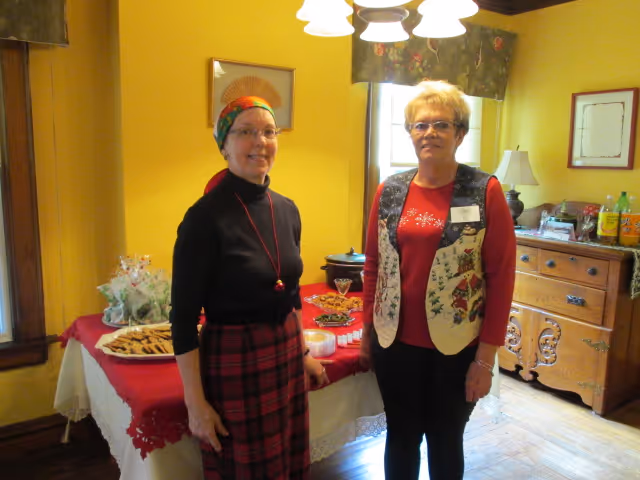 Two elderly women standing in a warmly lit room with yellow walls. One woman is wearing a black top, red plaid skirt, and a colorful headscarf, while the other is wearing glasses, a red shirt, and a festive vest. Behind them is a table covered with a red and white tablecloth, holding plates of cookies and other snacks. A wooden dresser with drinks and a lamp is visible in the background.